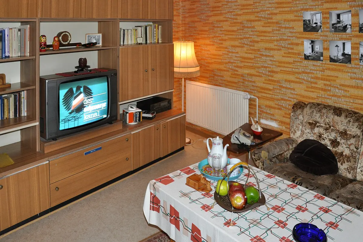 A GDR living room with a wooden wall unit, a television, patterned wallpaper, a telephone and a set table
